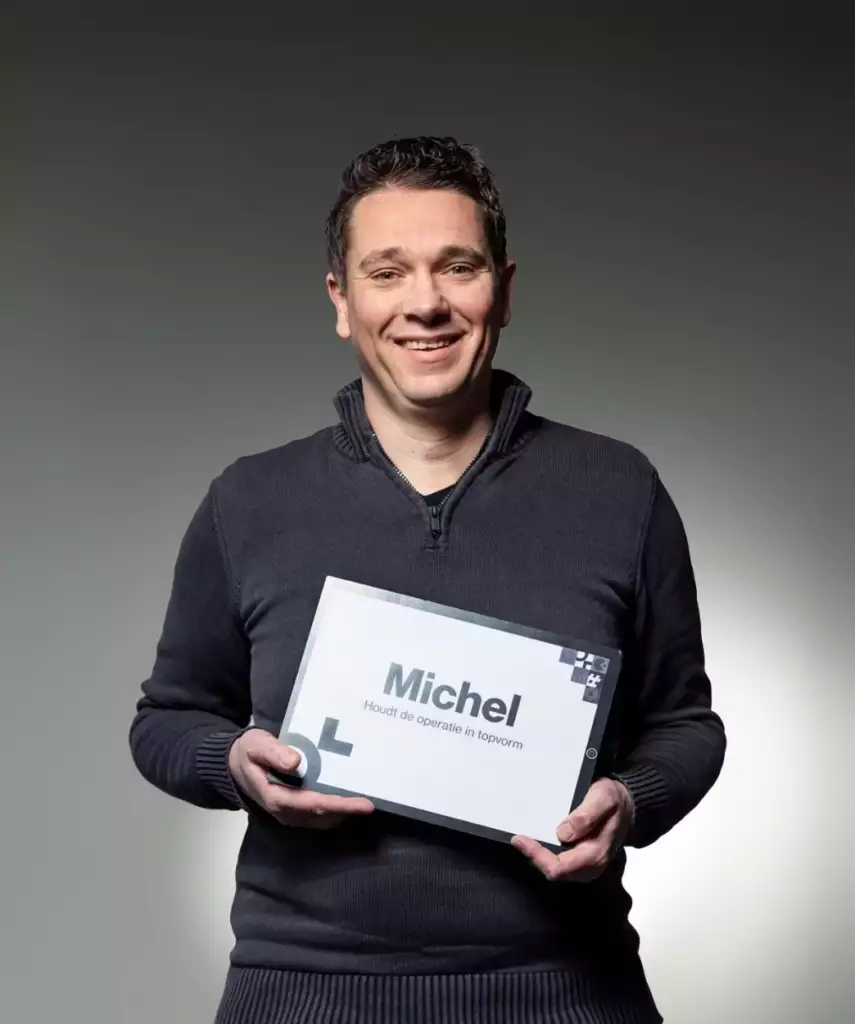 Een man met kort donker haar, draagt een donkere trui, glimlacht terwijl hij een bord met de naam Michel en Nederlandse tekst vasthoudt, staande tegen een effen, grijsverloop achtergrond.
