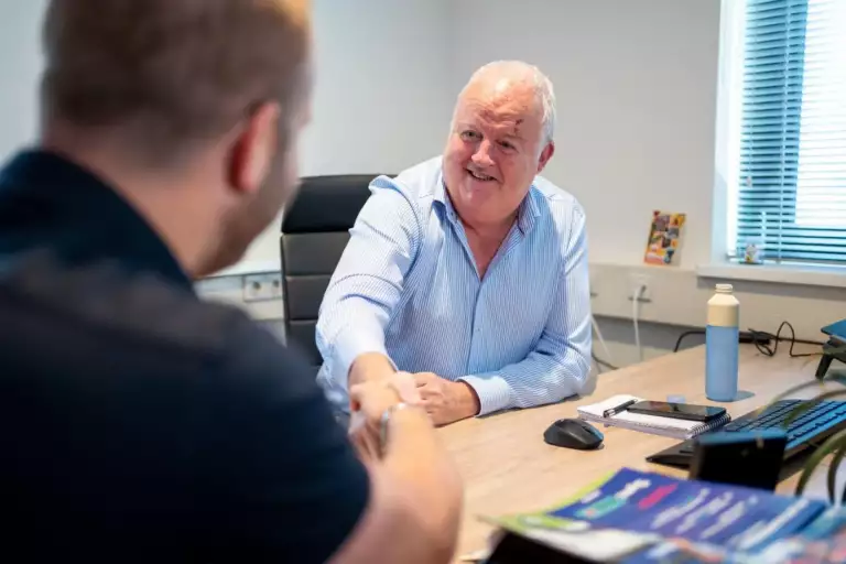 Twee mannen zitten tegenover elkaar aan een bureau in een kantoor en schudden elkaar de hand. De ene man, met grijs haar, glimlacht en draagt een lichtblauw overhemd. Op het bureau liggen papieren, een waterfles en kantoorbenodigdheden.