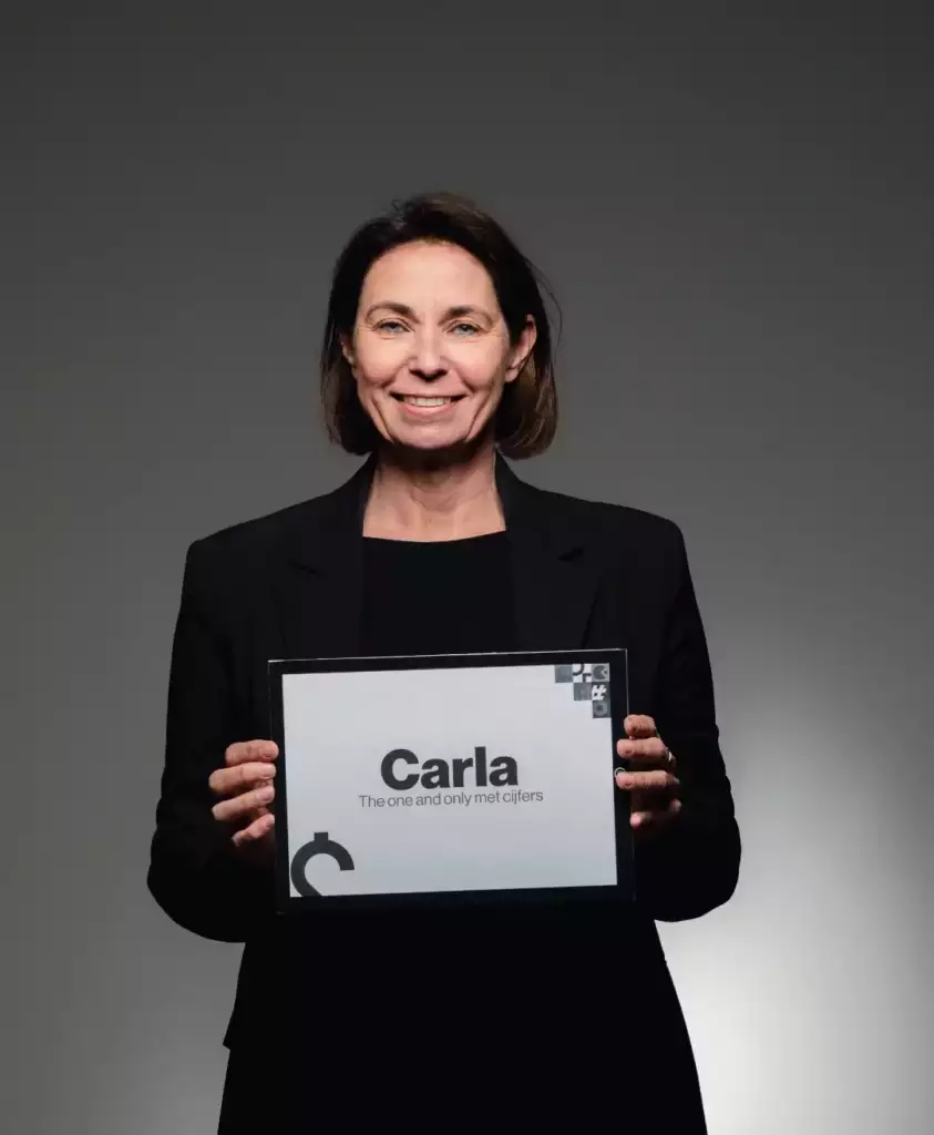 Een vrouw met schouderlang bruin haar, draagt een zwarte blazer, glimlacht terwijl ze een bord vasthoudt waarop staat, Carla: The one and only mct cjfers, tegen een effen grijze achtergrond.
