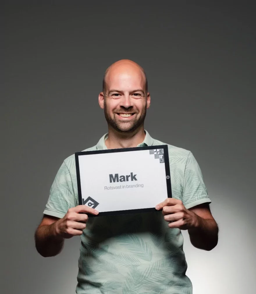 Een lachende man met een kaal hoofd en een lichte baard houdt een bord vast waarop staat: Mark, Rotsvast in branding. Hij draagt een lichtgroen poloshirt met patroon en staat tegen een effen grijze achtergrond.