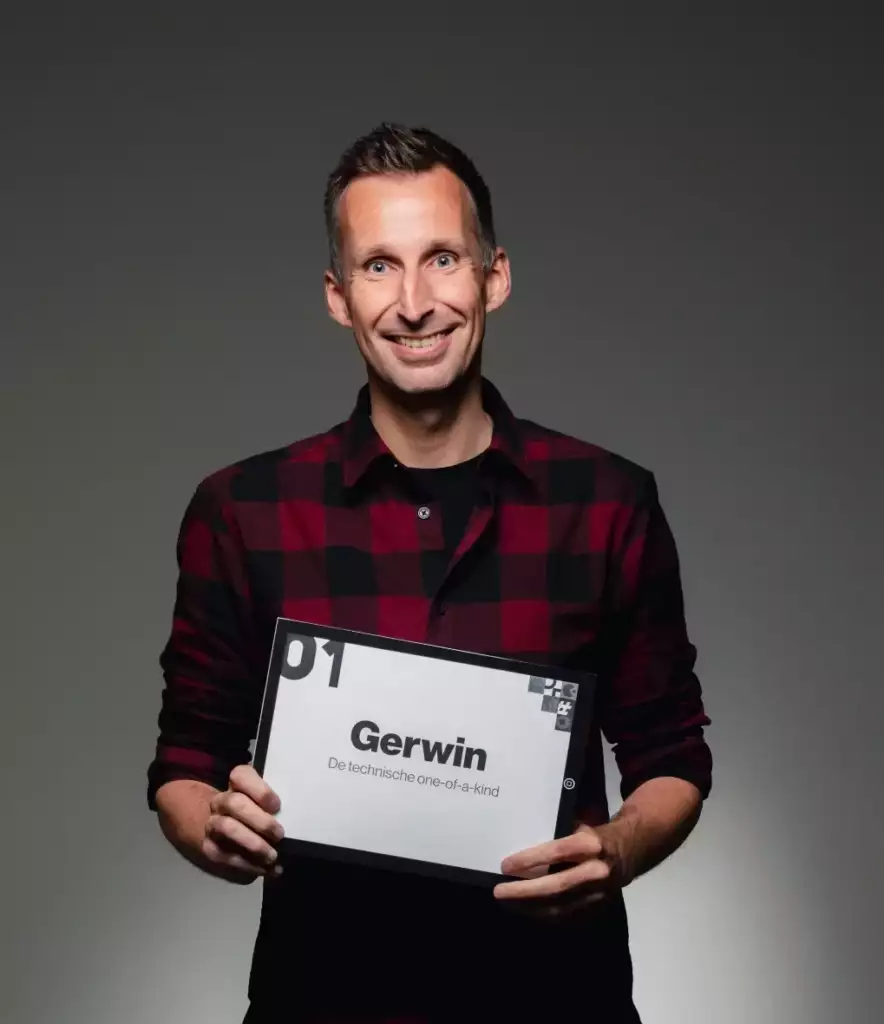 Een lachende man in een rood en zwart geruit overhemd houdt een bord vast waarop staat: 01 Gerwin De technische one-of-a-kind, tegen een effen, donkere achtergrond.