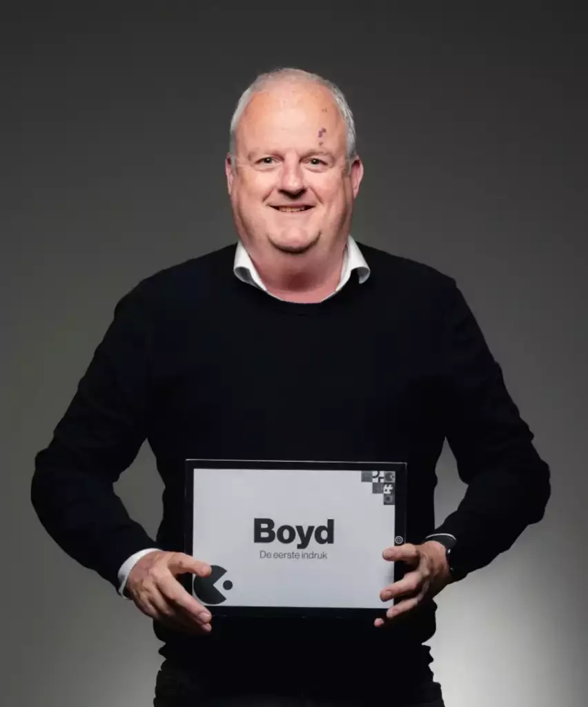 Een lachende man van middelbare leeftijd met grijs haar draagt een zwarte trui over een wit overhemd en houdt een bord vast waarop Boyd De eerste indruk staat tegen een effen, donkere achtergrond.