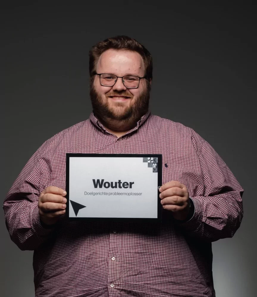Een man met een bril en een baard, in een rood geruit overhemd, glimlacht terwijl hij een bord vasthoudt waarop Wouter staat met kleinere tekst eronder tegen een donkere achtergrond.