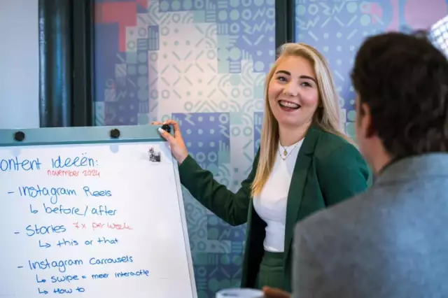 Een vrouw in een groen pak glimlacht terwijl ze inhoudelijke ideeën presenteert op een whiteboard aan een zittende collega in een kantoor met kleurrijke muren met patronen.