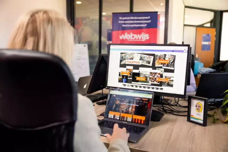 Een persoon met lang blond haar werkt aan een bureau met een laptop en een groot beeldscherm waarop websiteontwerpen te zien zijn. Op de achtergrond is een kantooromgeving met posters en glazen wanden zichtbaar.