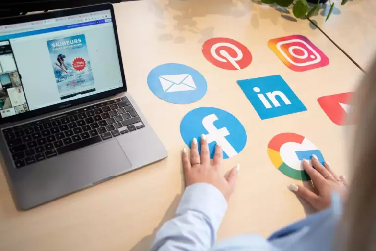 Een persoon zit achter een bureau met een laptop en verschillende grote sociale media-iconen, waaronder Facebook, LinkedIn, Pinterest, Instagram, YouTube, Google en een e-mailsymbool, weergegeven op het oppervlak.