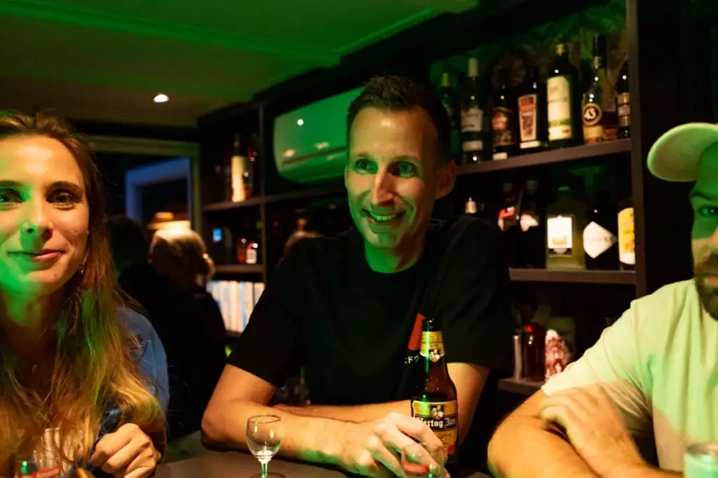 Drie mensen zitten aan een bartafel met drankjes, glimlachen en praten. Flessen staan op planken achter hen en de verlichting is schemerig, met een groenige tint op hun gezichten.