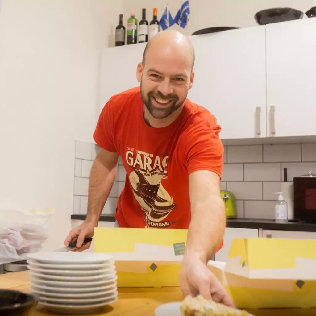 Een lachende man in een rood T-shirt serveert eten in een keuken, reikend over een aanrecht met gestapelde borden vlakbij en gele dozen voor hem. Op de achtergrond zijn flessen en keukengerei te zien.