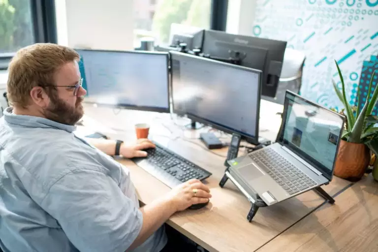 Een man met een bril werkt aan een bureau met drie computerschermen, een laptop, toetsenbord en muis. Op het bureau staan een potplant en een beker. Hij lijkt geconcentreerd op zijn schermen.
