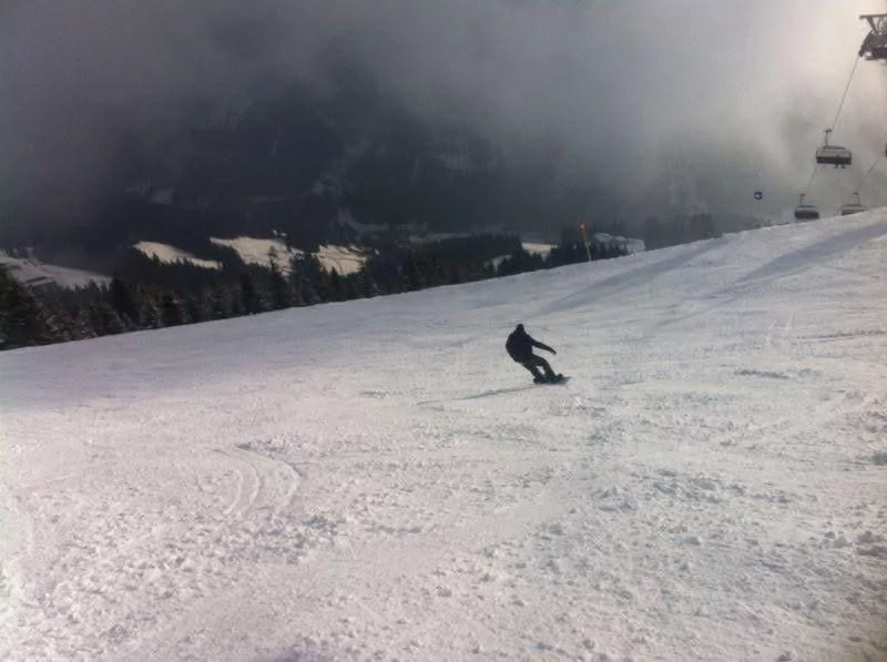 Een persoon snowboardt een brede, besneeuwde helling af met bomen en skiliften zichtbaar op de achtergrond onder een bewolkte hemel.