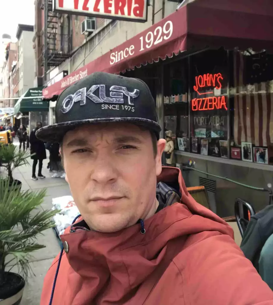 Een man in een rode jas en een zwarte Oakley pet neemt een selfie voor John's Pizzeria in een straat in de stad. Het uithangbord en de luifel van het restaurant en andere voetgangers zijn zichtbaar op de achtergrond.