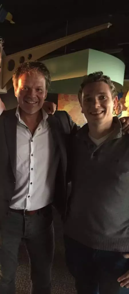 Twee mannen staan binnen naast elkaar en glimlachen naar de camera. De ene draagt een colbert en wit overhemd, terwijl de andere gekleed is in een grijze trui. De achtergrond is schemerig verlicht met een kleurrijk decor.