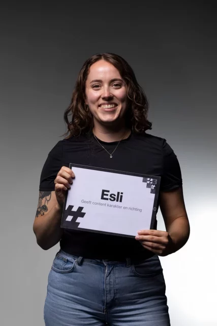 Een glimlachende persoon met lang bruin haar houdt een bordje vast met de tekst „Esli Geeft inhoud, karakter en richting”, tegen een grijze achtergrond met kleurverloop. De persoon, gekleed in een zwart shirt en een blauwe spijkerbroek, belichaamt de creativiteit en strategie die vaak in online marketing te vinden zijn.