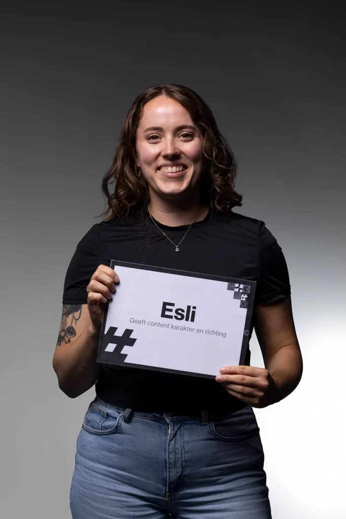 Een glimlachende persoon met lang bruin haar houdt een bordje vast met de tekst „Esli Geeft inhoud, karakter en richting”, tegen een grijze achtergrond met kleurverloop. De persoon, gekleed in een zwart shirt en een blauwe spijkerbroek, belichaamt de creativiteit en strategie die vaak in online marketing te vinden zijn.