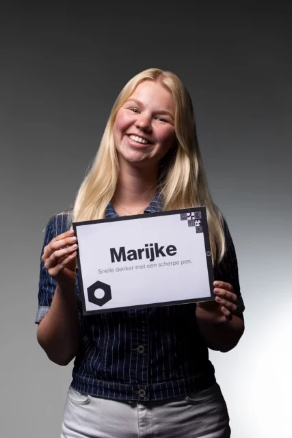 Een glimlachende jonge vrouw met lang blond haar houdt een bordje vast waarop staat: „Marijke Snelle, denker met een scherpe pen”. De achtergrond is grijs en de belichting benadrukt haar gezicht, waardoor ze de zelfverzekerde uitstraling heeft van een expert op het gebied van online marketing.