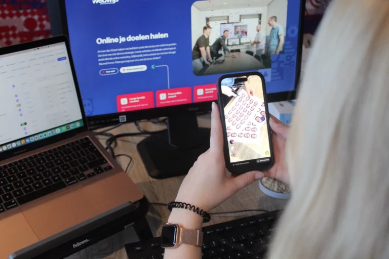 Een persoon met een smartwatch is bezig op haar telefoon terwijl ze de website van Webwijs open heeft staan op het scherm erachter