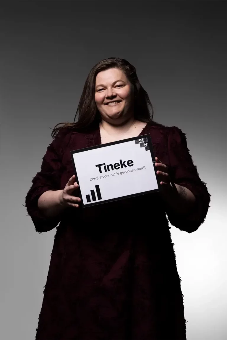 Een glimlachende vrouw met lang bruin haar, die een jurk met donkere structuur draagt, houdt een wit bord vast waarop staat: Tineke. Zorgt ervoor dat jij gehoord wordt tegen een grijsverloop achtergrond.