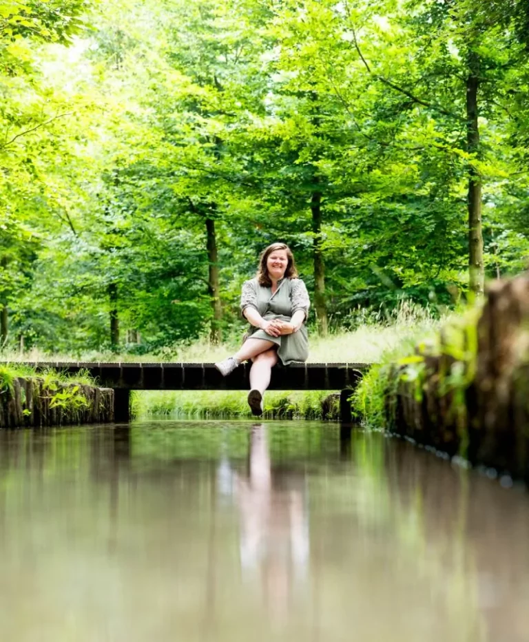 Tineke zit op een kleine houten brug over een kalm beekje, omringd door weelderige groene bomen en gras op een heldere, zonnige dag.