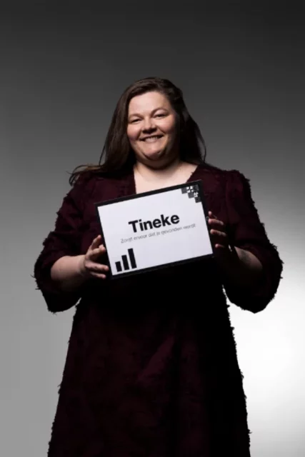 Een glimlachende vrouw met lang bruin haar in een bordeauxrode jurk houdt een bord vast waarop staat: Tineke. Zorgt ervoor dat je gevonden wordt, met drie zwarte staafdiagrammen onder de tekst. De achtergrond is grijs.