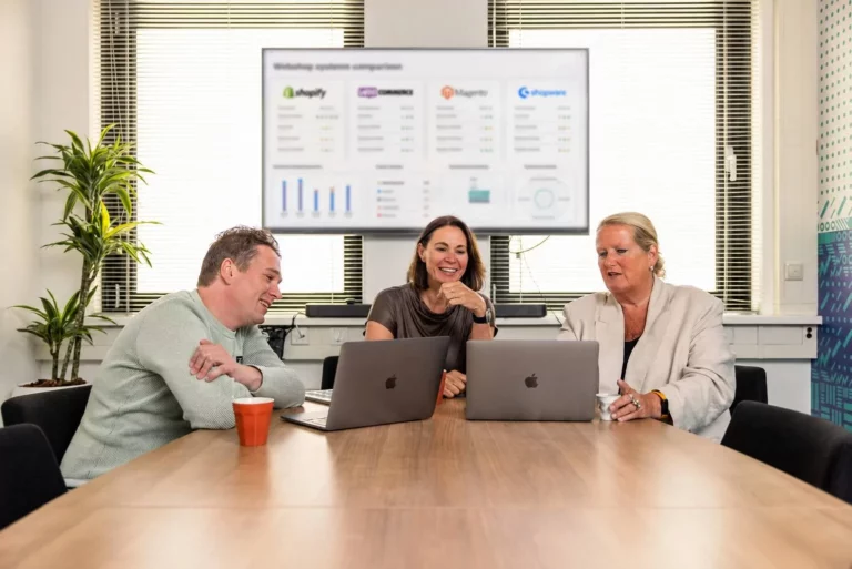 Drie mensen zitten aan een vergadertafel met hun laptops, glimlachend en in gesprek. Op een scherm achter hen is een vergelijking van webshopsystemen te zien, met grafieken en logo’s.