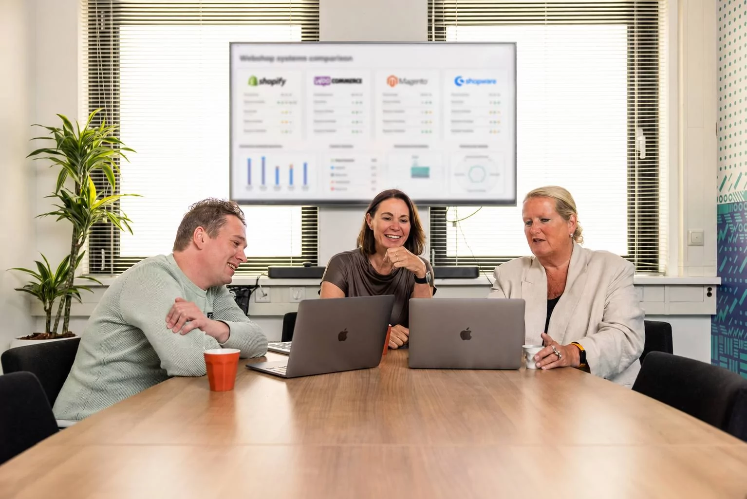 Drie mensen zitten aan een vergadertafel met hun laptops, glimlachend en in gesprek. Op een scherm achter hen is een vergelijking van webshopsystemen te zien, met grafieken en logo’s.