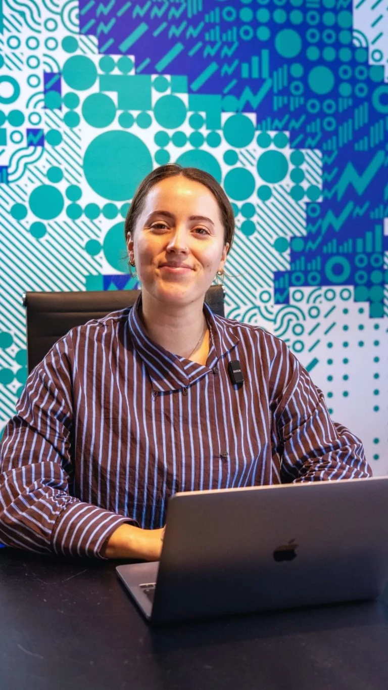 Een vrouw in een gestreept Meta One-shirt zit aan een bureau met een laptop voor zich, tegen een muur met een blauw-wit patroon.