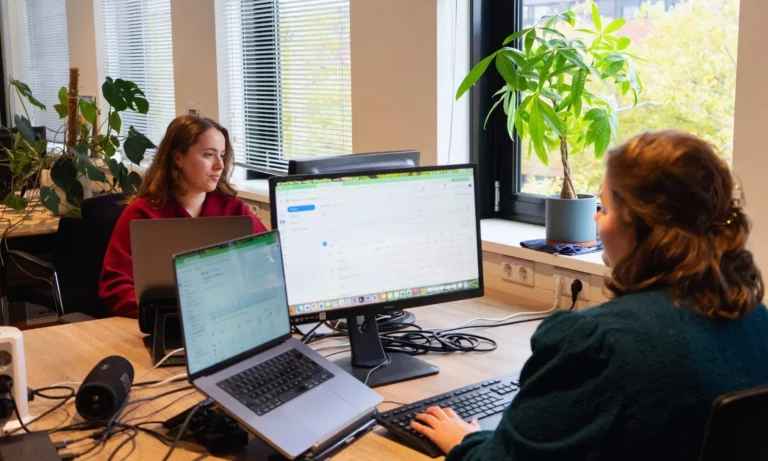 Twee vrouwen die aan een gedeeld bureau werken op laptops en een Meta One-desktopmonitor.